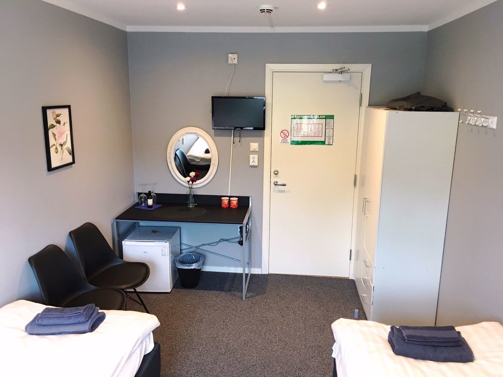 Une chambre d’hôtel de taille modeste, équipée de deux lits, d’un bureau avec un miroir, d’une télévision et d’une porte sur laquelle est apposé un panneau « interdiction de fumer ».