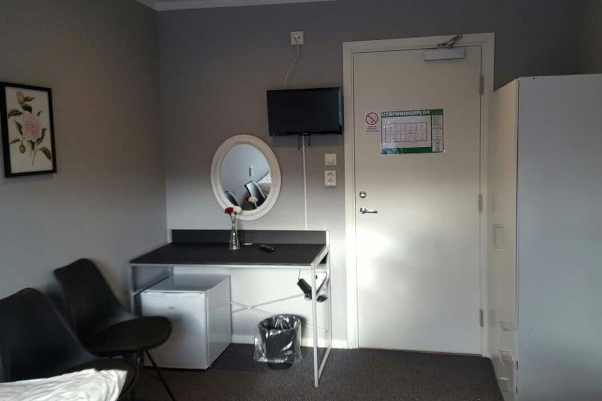 Ein kleines Hotelzimmer mit einem Bett, einem Schreibtisch inklusive Spiegel, einem Fernseher, einem Kühlschrank und einer Mülleimer.