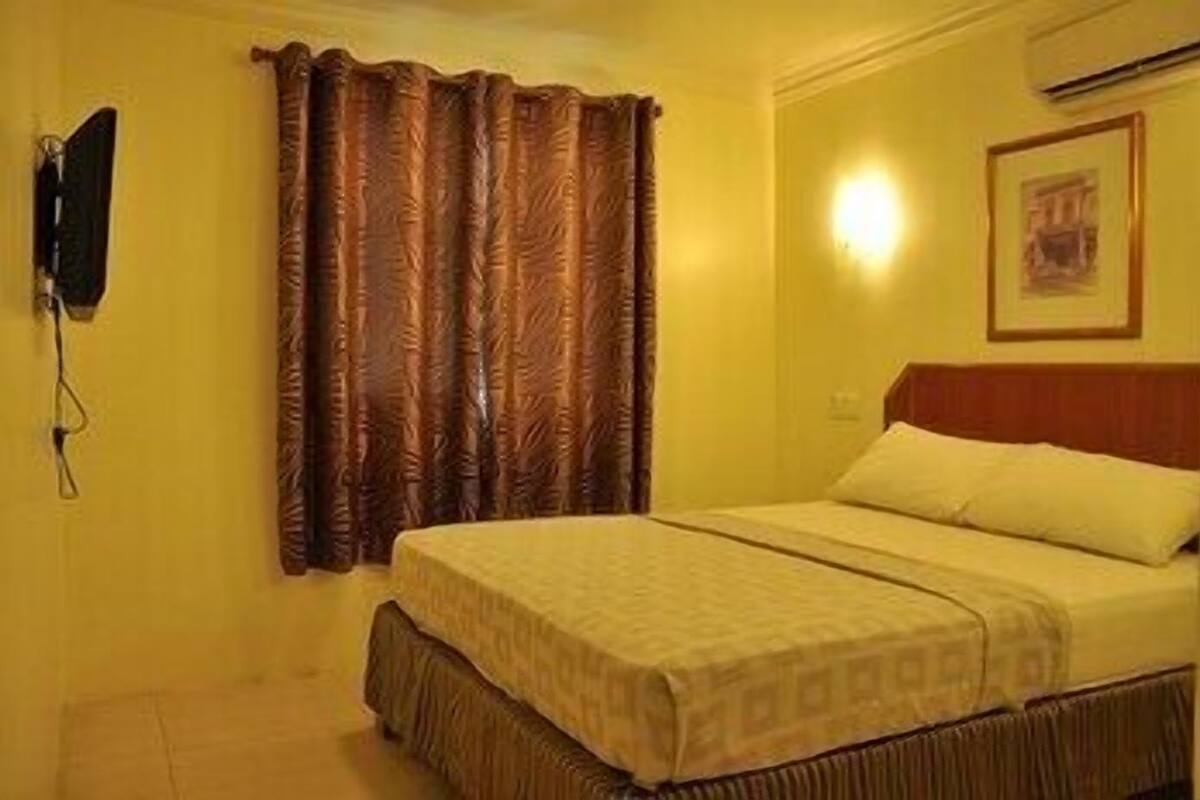 Una habitación de hotel con cama, cortinas, un televisor montado en la pared y un cuadro enmarcado.