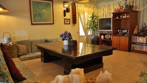 A living room with a large dark wooden coffee table, a sofa with patterned cushions, a bookshelf filled with books and decorative items, and a television on a wooden stand. - Johan Travellers Lodge (Malacca City)