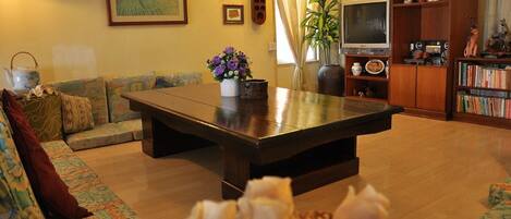 Un salón con una amplia mesa de centro de madera oscura, un sofá con cojines estampados, una estantería llena de libros y objetos decorativos, y un televisor sobre un mueble de madera.