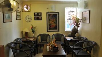 A dining area with wicker chairs, a wooden table, and a vase with flowers. There are framed pictures on the walls and a clock.