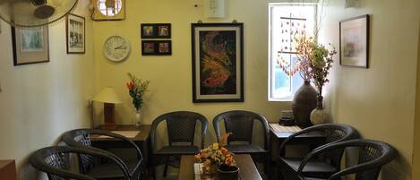 A dining area with wicker chairs, a wooden table, and a vase with flowers. There are framed pictures on the walls and a clock.