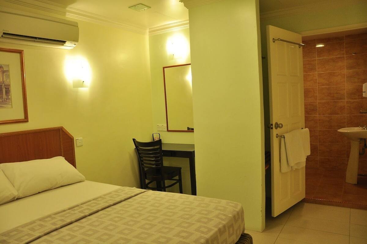 Habitación de hotel con cama, escritorio, silla, espejo y baño con lavabo y ducha.