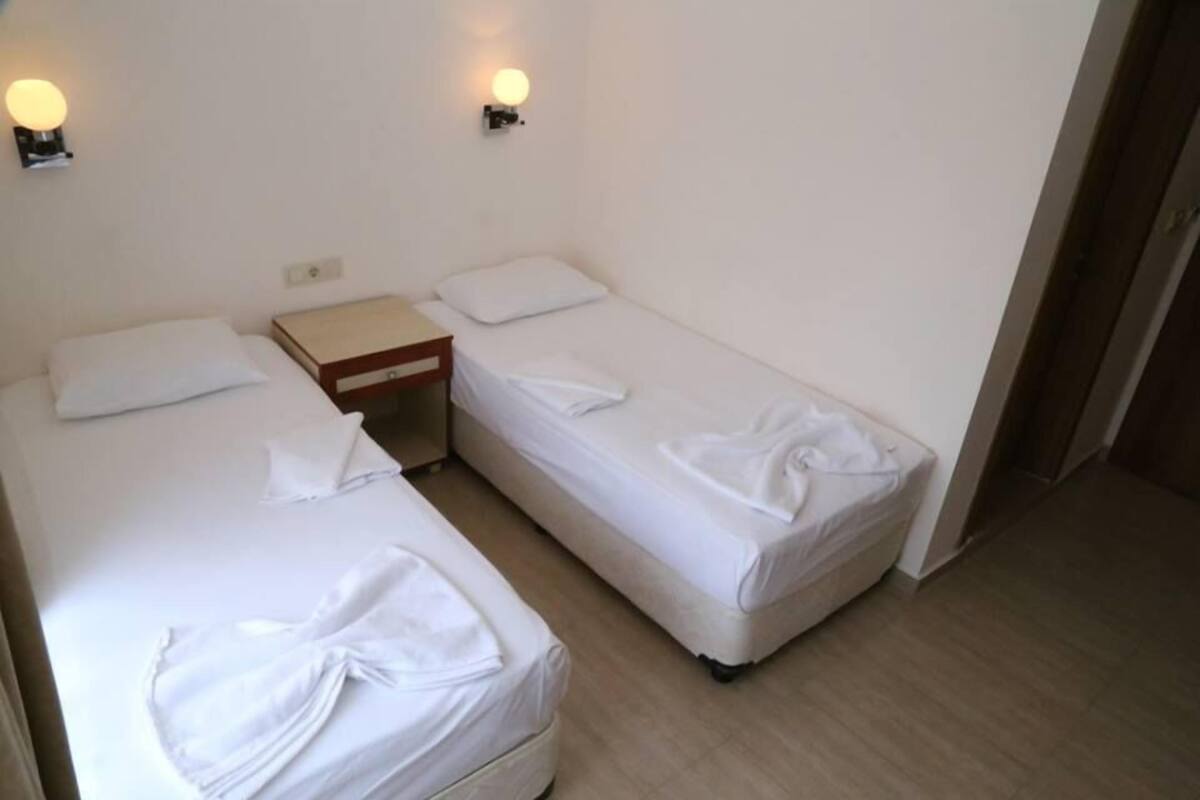 Una habitación con dos camas individuales, cada una con una almohada y una manta, una mesita de noche de madera y dos lámparas fijadas a la pared.