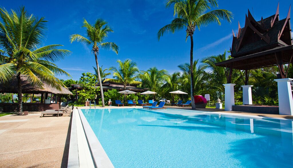 A swimming pool with lounge chairs and umbrellas, surrounded by palm trees and a traditional building.