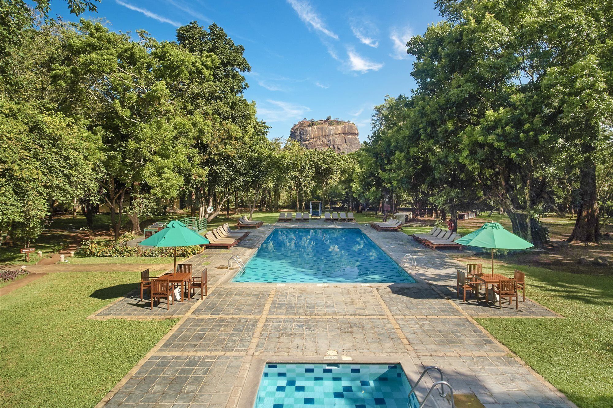 A rectangular swimming pool with a stone border, surrounded by lounge chairs and umbrellas, with trees and a distant rock formation.