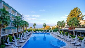 Una piscina con sillones de playa y sombrillas, rodeada de palmeras y un edificio de hotel.