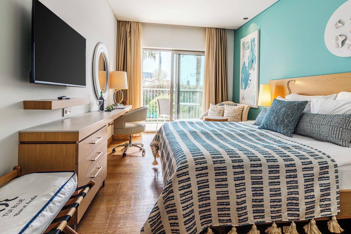 A hotel room with a bed, a desk, a chair, a TV, a window with curtains, and a balcony.