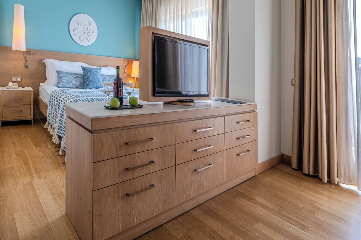 A modern hotel room with a wooden cabinet, a bed with blue bedding, a TV on a stand, and a view of the city through large windows.