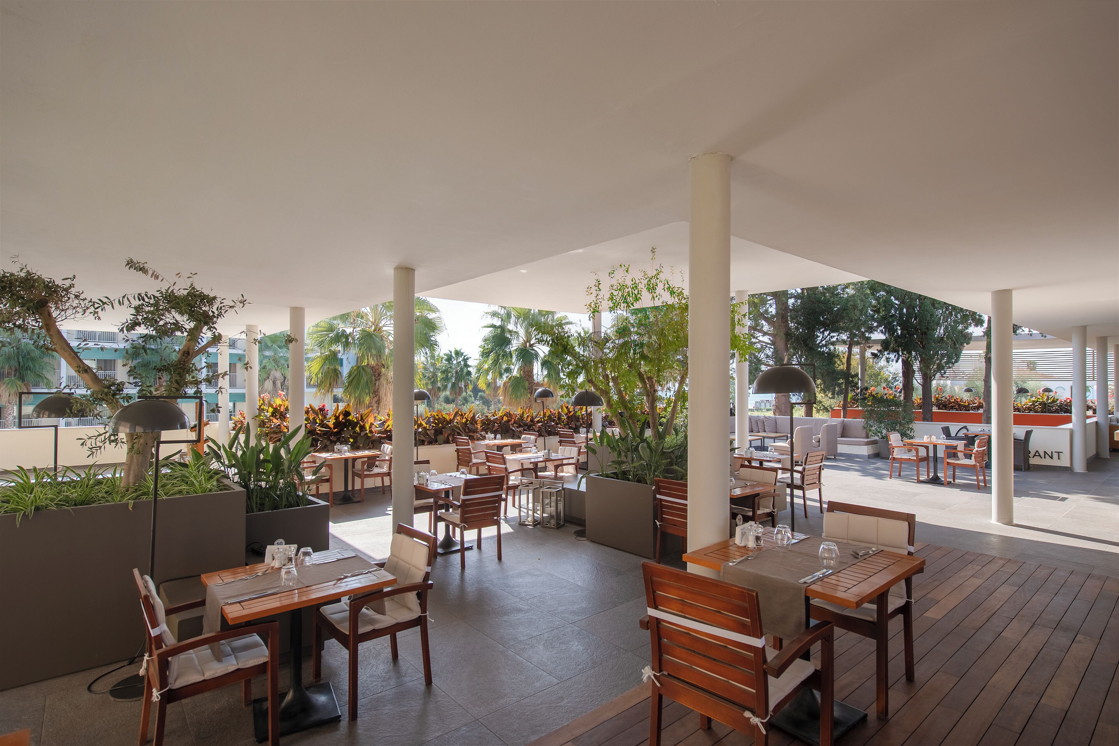Un área de comedor al aire libre con mesas y sillas de madera, rodeada de macetas y columnas.
