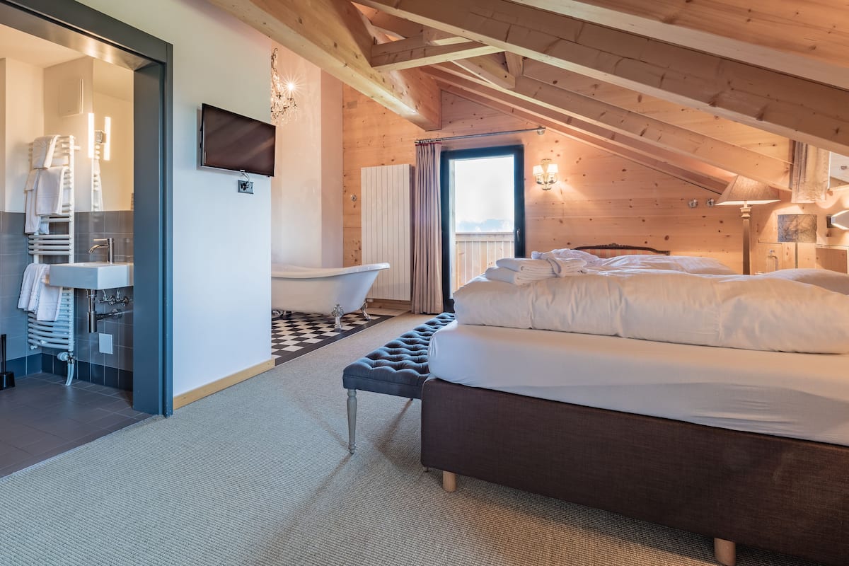A bedroom with a bed, a bench, a bathtub, and a bathroom with a sink and towel rack.