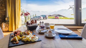 A breakfast table with a variety of foods, a window view of a mountain and a parked car, and a wooden table.