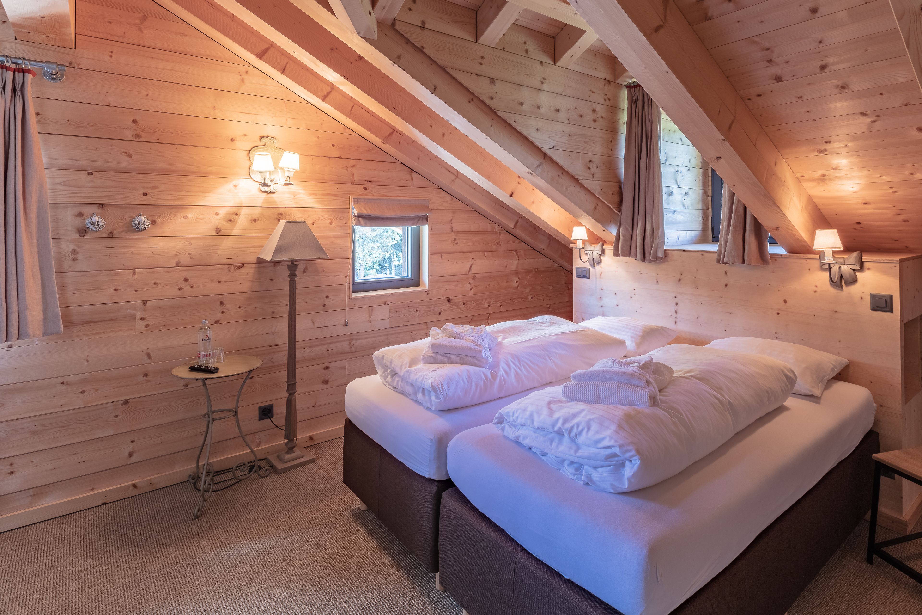 Une pièce chaleureuse aux murs en bois, avec un lit, deux lampes de chevet, une petite fenêtre et une table de chevet surmontée d’une lampe.
