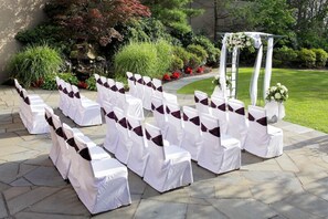 Un aménagement extérieur formel avec des chaises blanches et des rubans foncés, une zone pavée de pierres et une arche ornée de fleurs.
