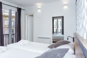 Une chambre d’hôtel avec un lit, deux oreillers, une fenêtre avec des rideaux, un radiateur et un miroir qui reflète la pièce.