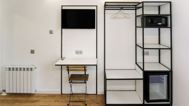 Ein Zimmer mit weißem Schreibtisch, einem Sessel, einem an der Wand montierten schwarzen Fernseher und einer schwarzen Regaleinheit mit Kleiderstange.