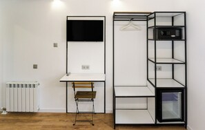 A room with a white desk, a chair, a black TV mounted on the wall, and a black shelving unit with a hanging clothes rack. - Fontanella Green House (Barcelona)