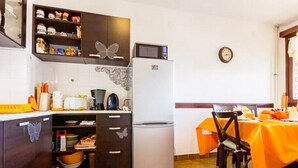 A compact kitchen with a refrigerator, microwave, and various kitchen appliances. There is a dining table with an orange tablecloth and chairs. The kitchen has dark wooden cabinets with decorative butterfly cutouts. - Apartment Palcic (Zadar)