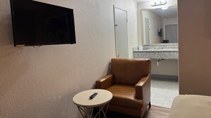 A hotel room with a television mounted on the wall, a brown leather chair, a small round table with a remote control, and a bathroom visible through an open door. - Harmony Inn and Suites (Beaumont)