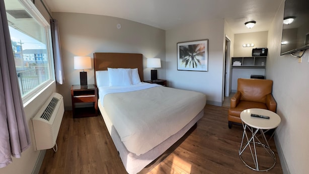 A hotel room with a large bed, two bedside tables with lamps, a brown leather chair, a small round table with a remote, and a TV on the wall. - Harmony Inn and Suites (Beaumont)