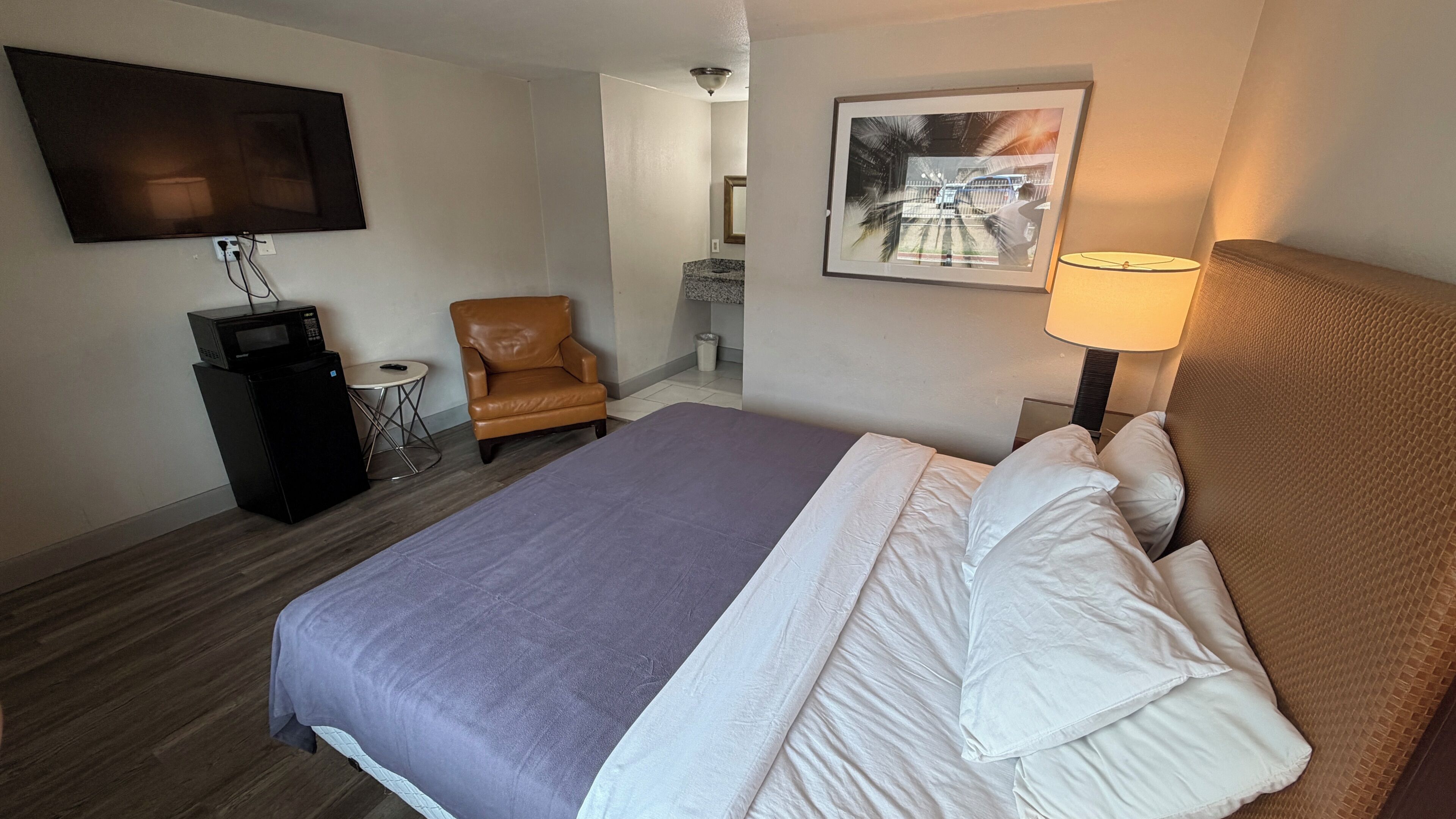 A hotel room with a bed, a television, a chair, a desk with a lamp, and a framed picture on the wall.
