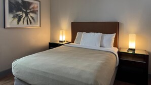 A hotel room with a bed, two bedside tables with lamps, a framed picture on the wall, and a smoke detector on the ceiling. - Harmony Inn and Suites (Beaumont)