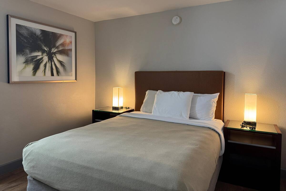 A hotel room with a bed, two bedside tables with lamps, a framed picture on the wall, and a smoke detector on the ceiling.