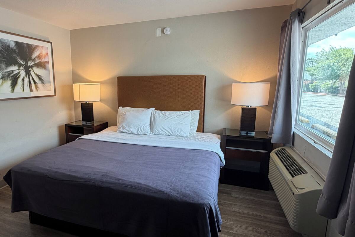 A hotel room with a bed, two bedside tables with lamps, a wall-mounted air conditioner, and a window with curtains.