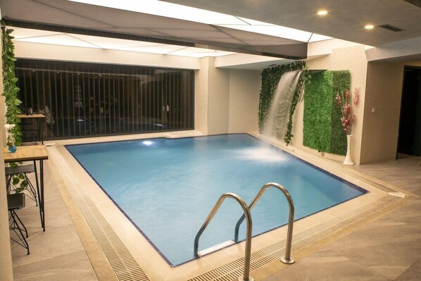 An indoor swimming pool with a waterfall feature, surrounded by a tiled floor and a glass partition.
