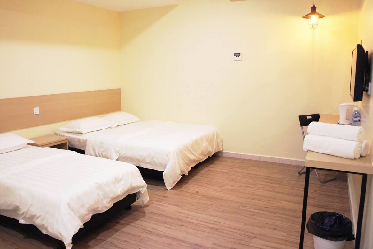 A hotel room with two beds, a wooden floor, a desk with a trash bin, and a wall-mounted light fixture.