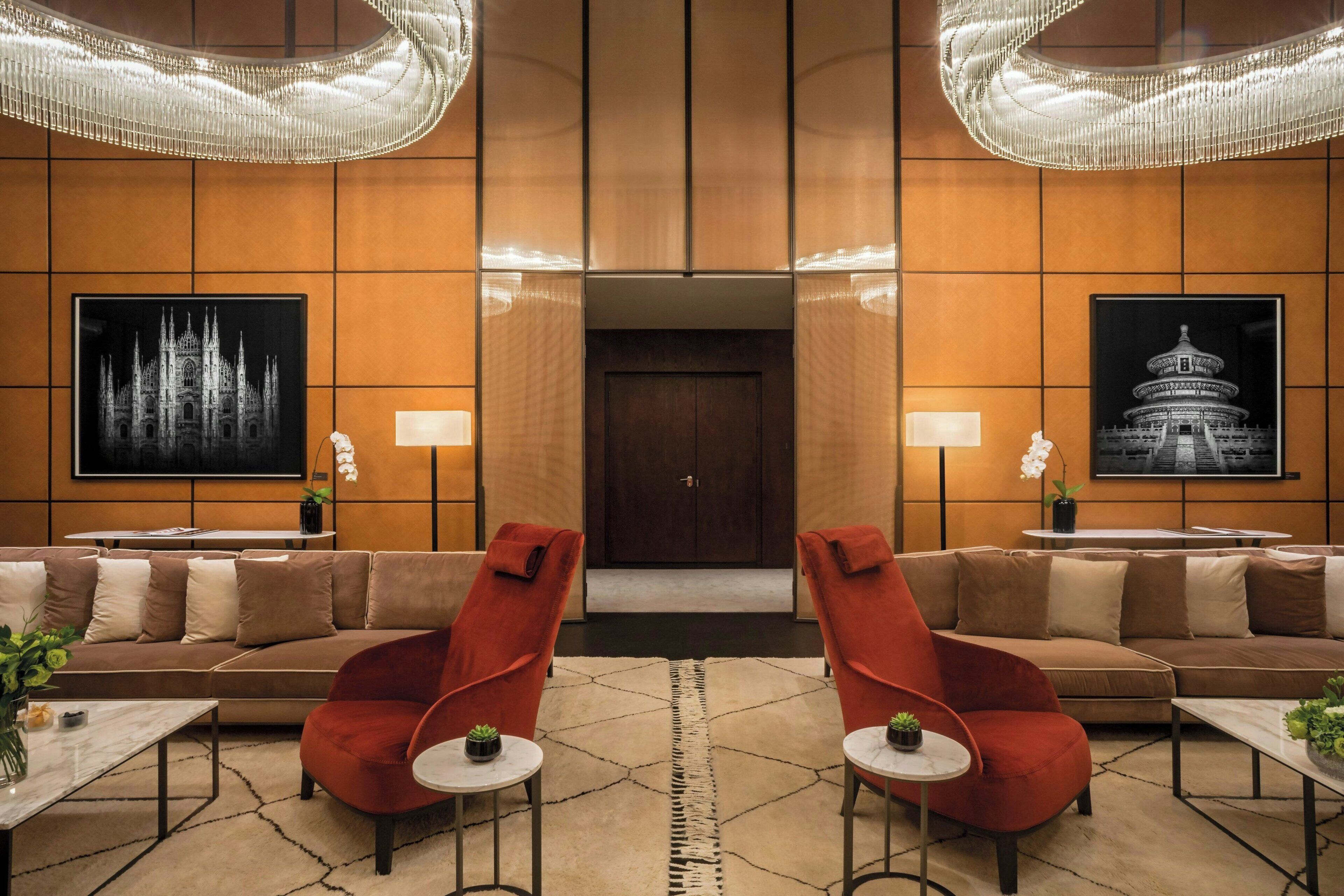 Un hall d’entrée avec un canapé marron, des fauteuils rouges, un lustre et deux tableaux encadrés.