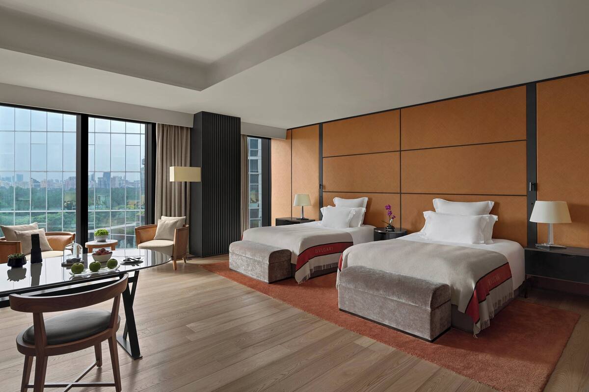 A hotel room with two beds, a dining table with chairs, a view of the city, and a large window.