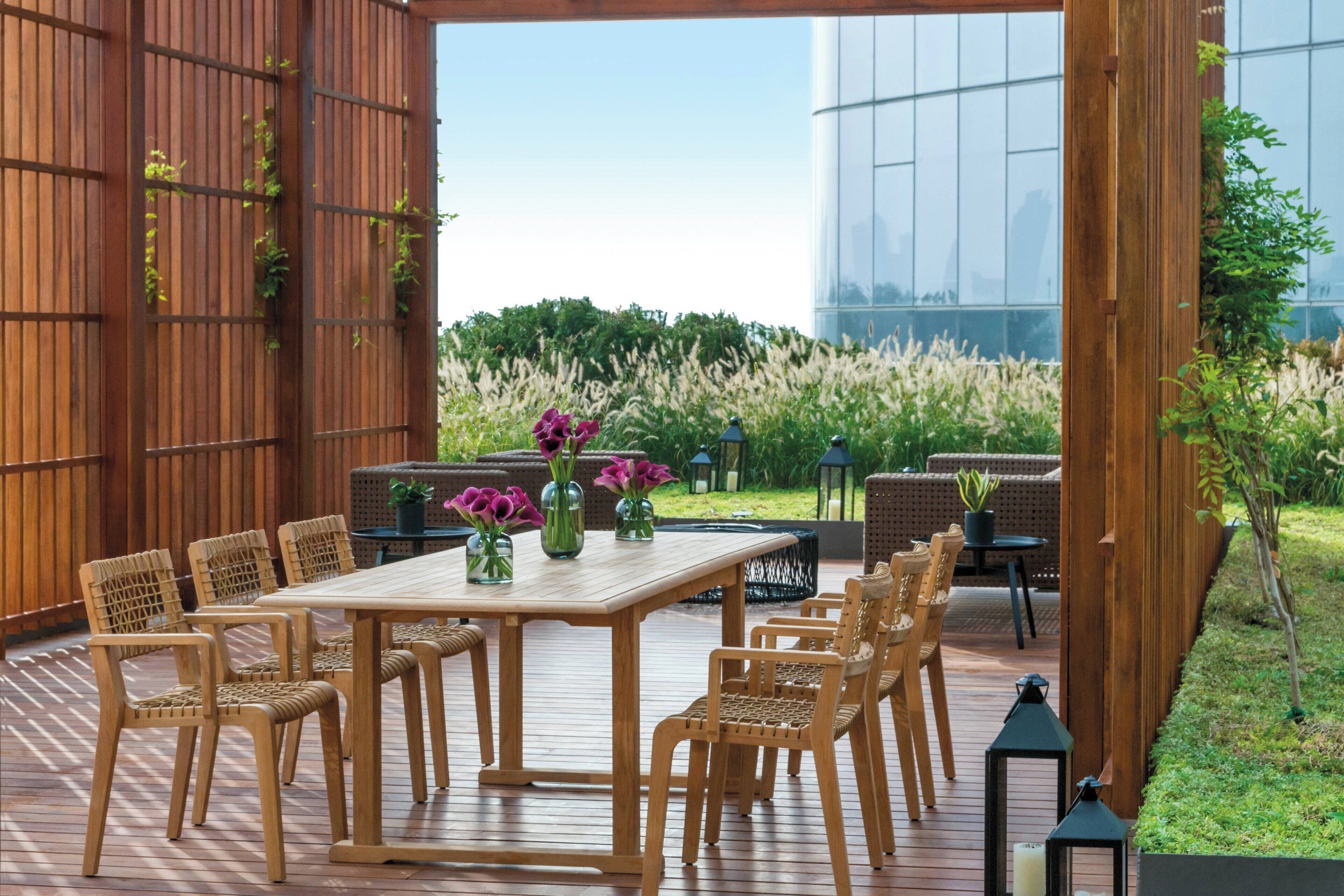 Un patio al aire libre con una mesa y sillas de madera, rodeado de abundante vegetación y vistas a un edificio moderno.