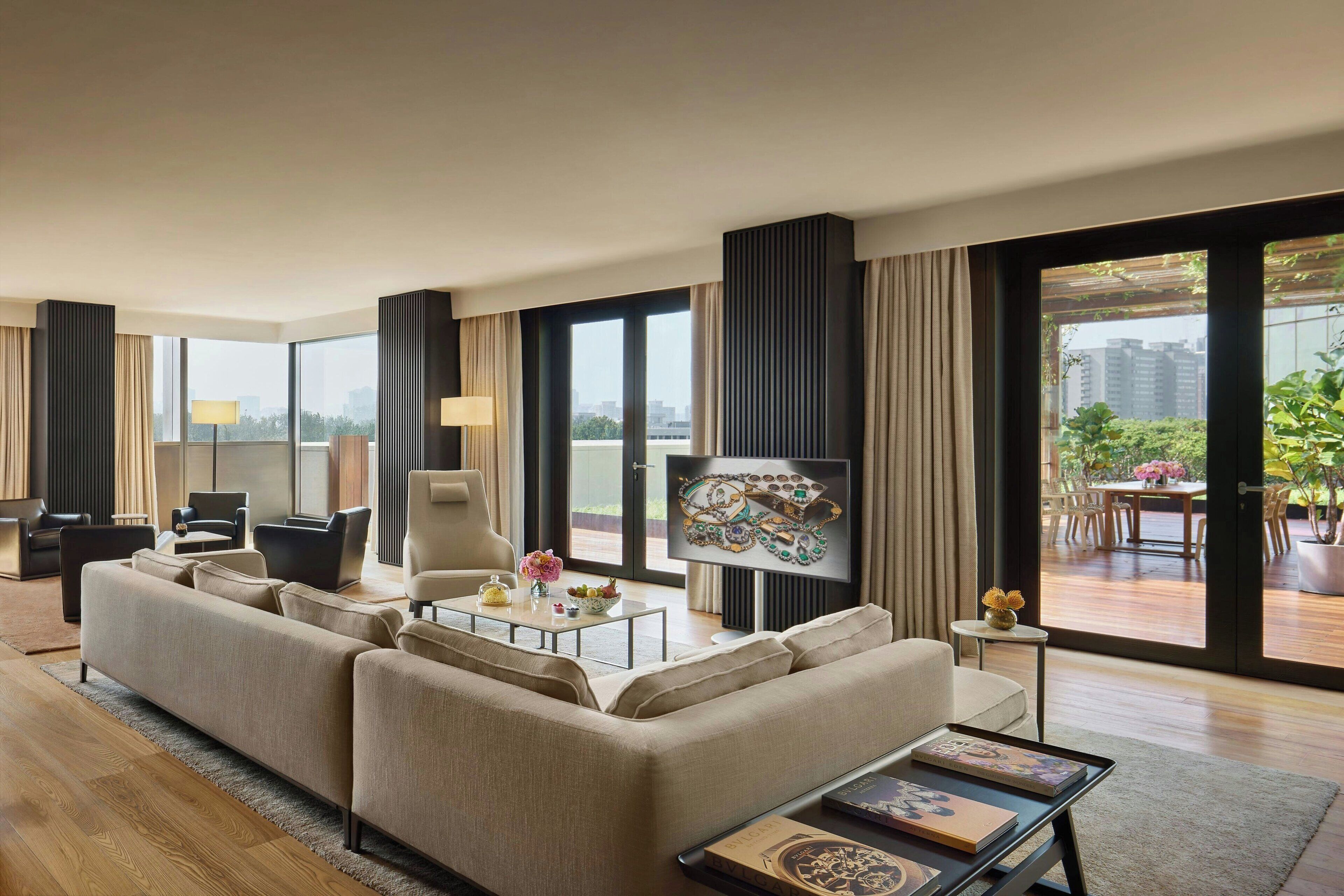 Una sala de estar moderna con sofá, mesa de centro y televisor. Hay cortinas, una lámpara y vistas al exterior.