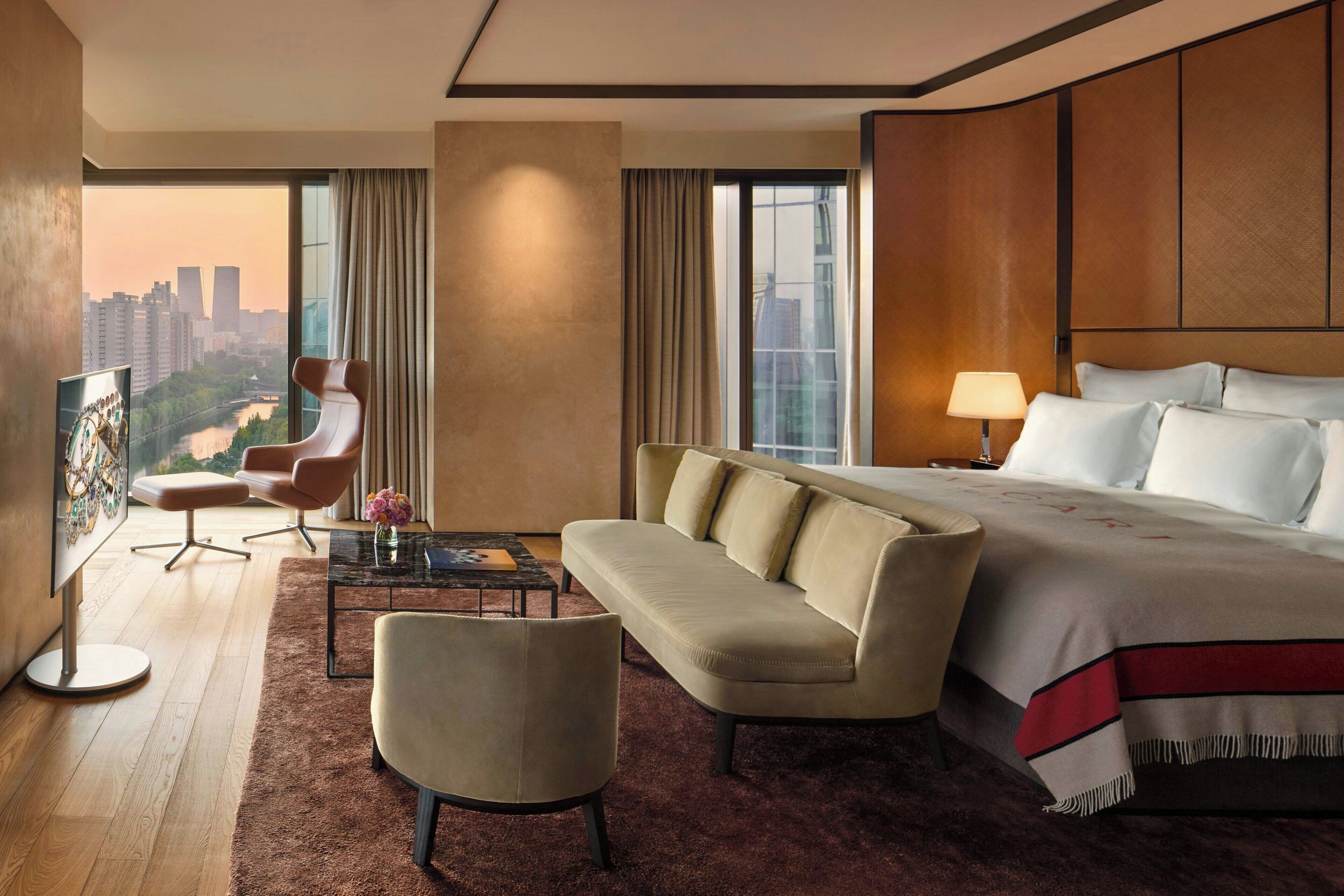 A hotel room with a large bed, a sofa, a desk with a lamp, a chair, and a view of a cityscape through large windows.