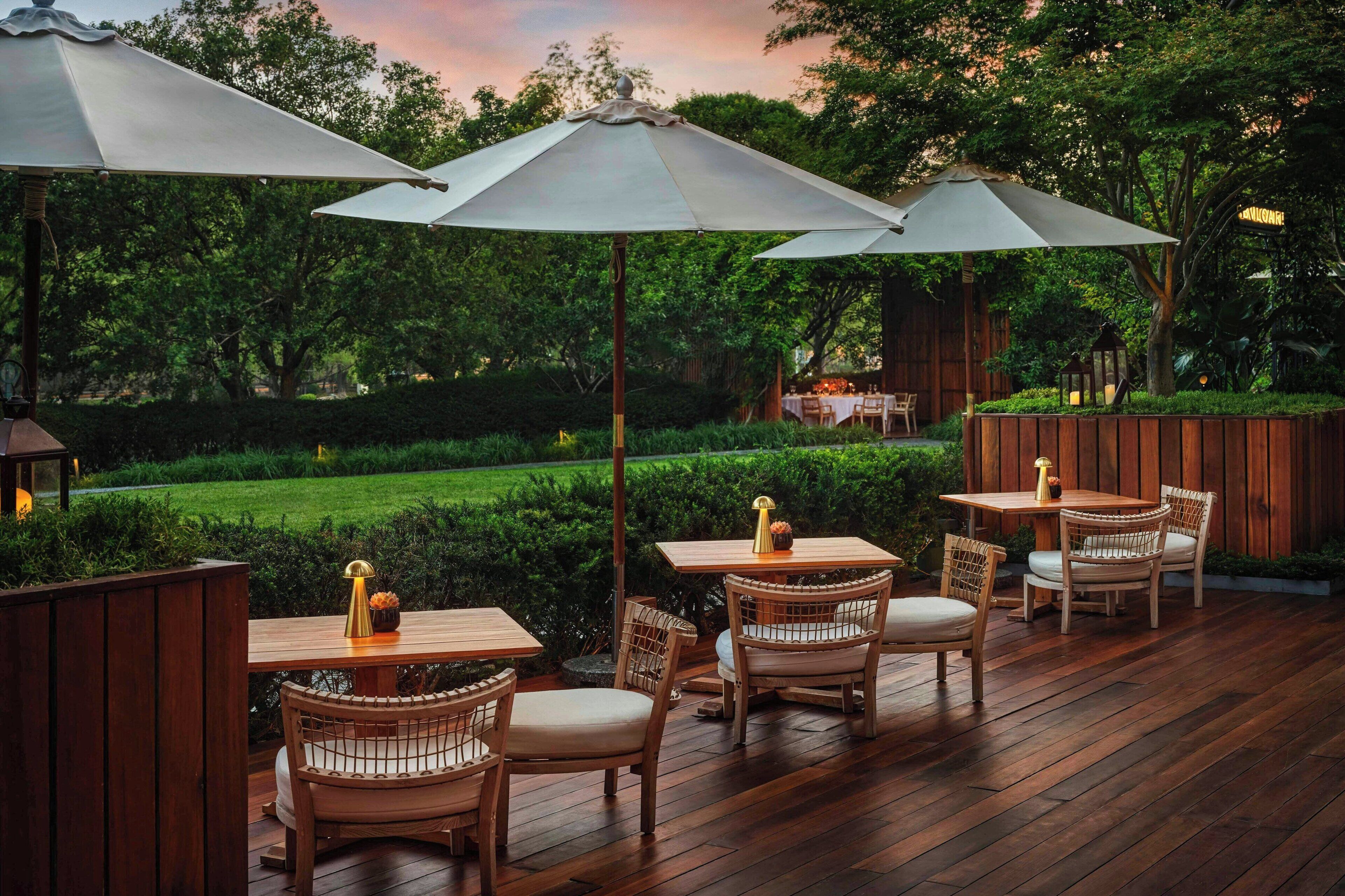 Eine Terrasse im Freien mit Holztischen und -stühlen, umgeben von üppiger Bepflanzung und Bäumen.