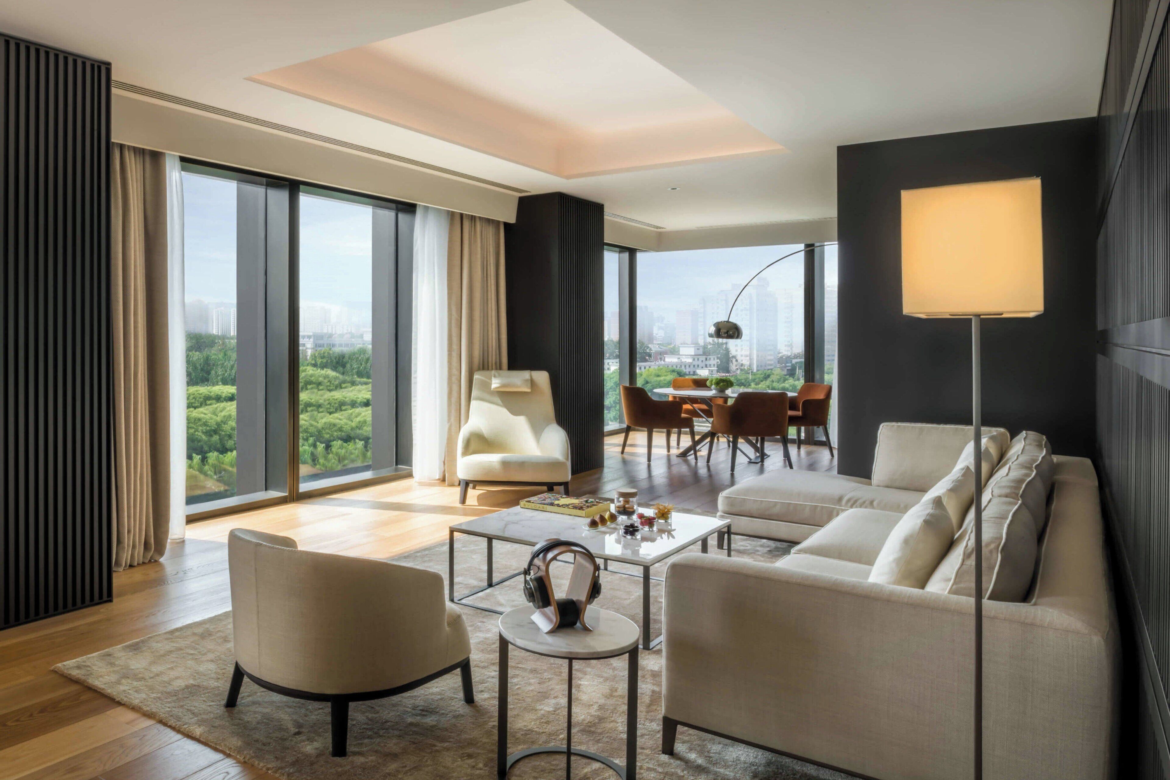 Un salón moderno con un sofá, sillones, una mesa de centro y una lámpara de pie. Una ventana grande ofrece vistas a zonas verdes y edificios.