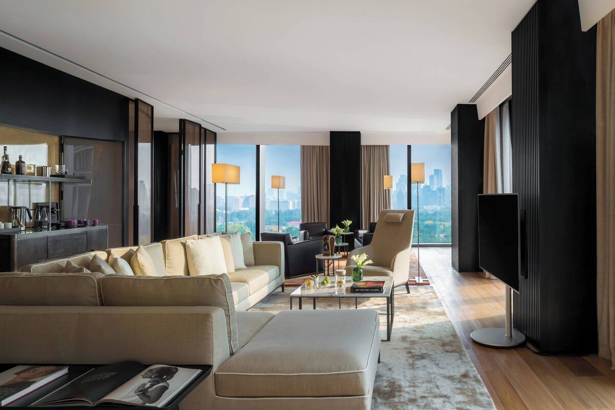 A modern living room with a sofa, ottoman, and coffee table, a flat-screen TV, and large windows with a city view.