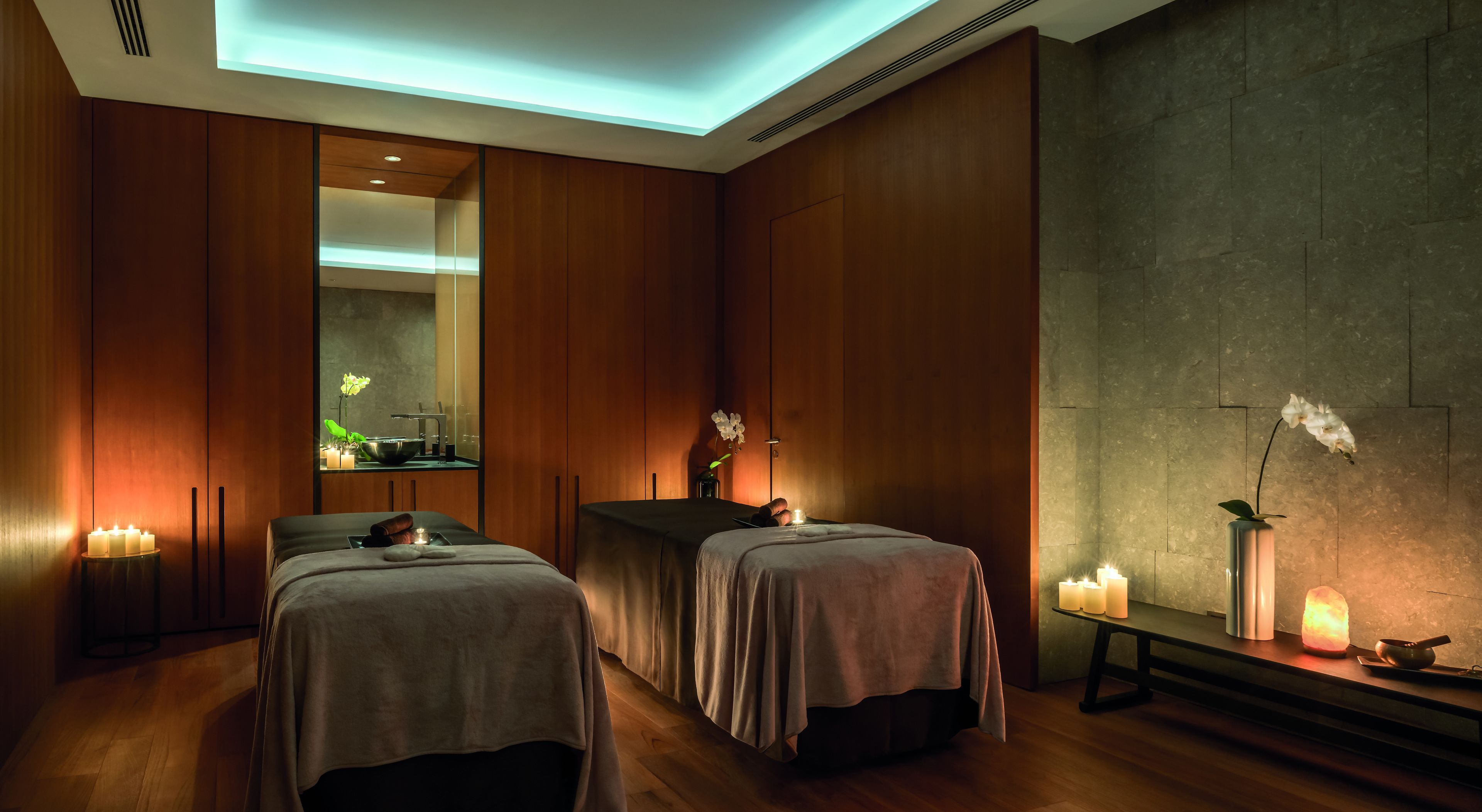 Een spa-ruimte met twee massagetafels, kaarsen en een spiegel die een wastafel en groen reflecteert.
