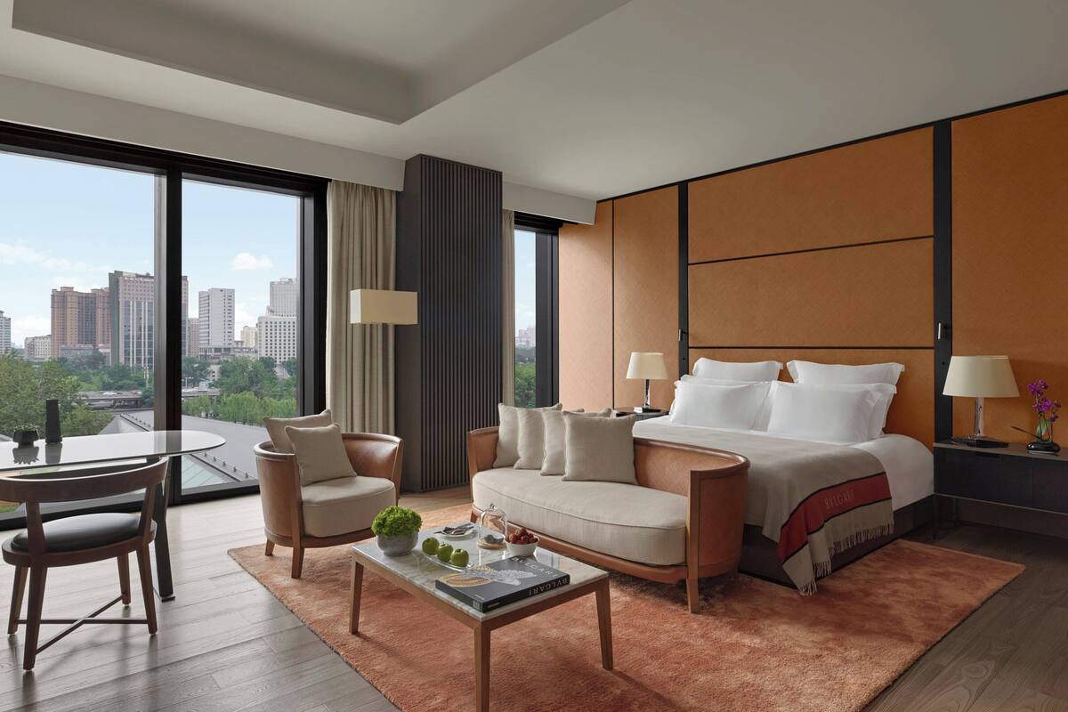 A modern hotel room with a large bed, a sofa, a dining table, and a view of the cityscape.