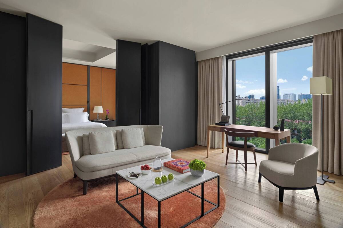 A modern hotel room with a sofa, a coffee table, a desk, and a large window offering a city view.