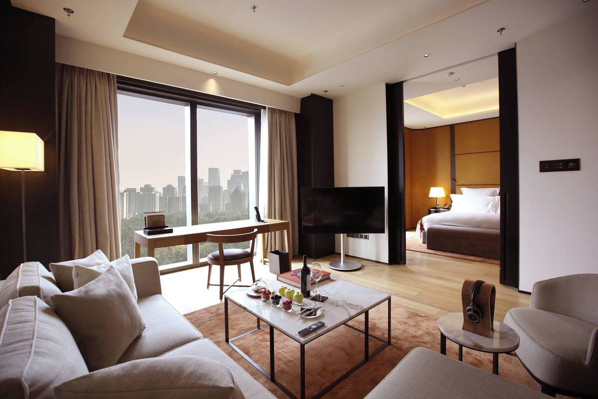 A hotel room with a large window, a sofa, a coffee table, a TV, a bed, and a desk.
