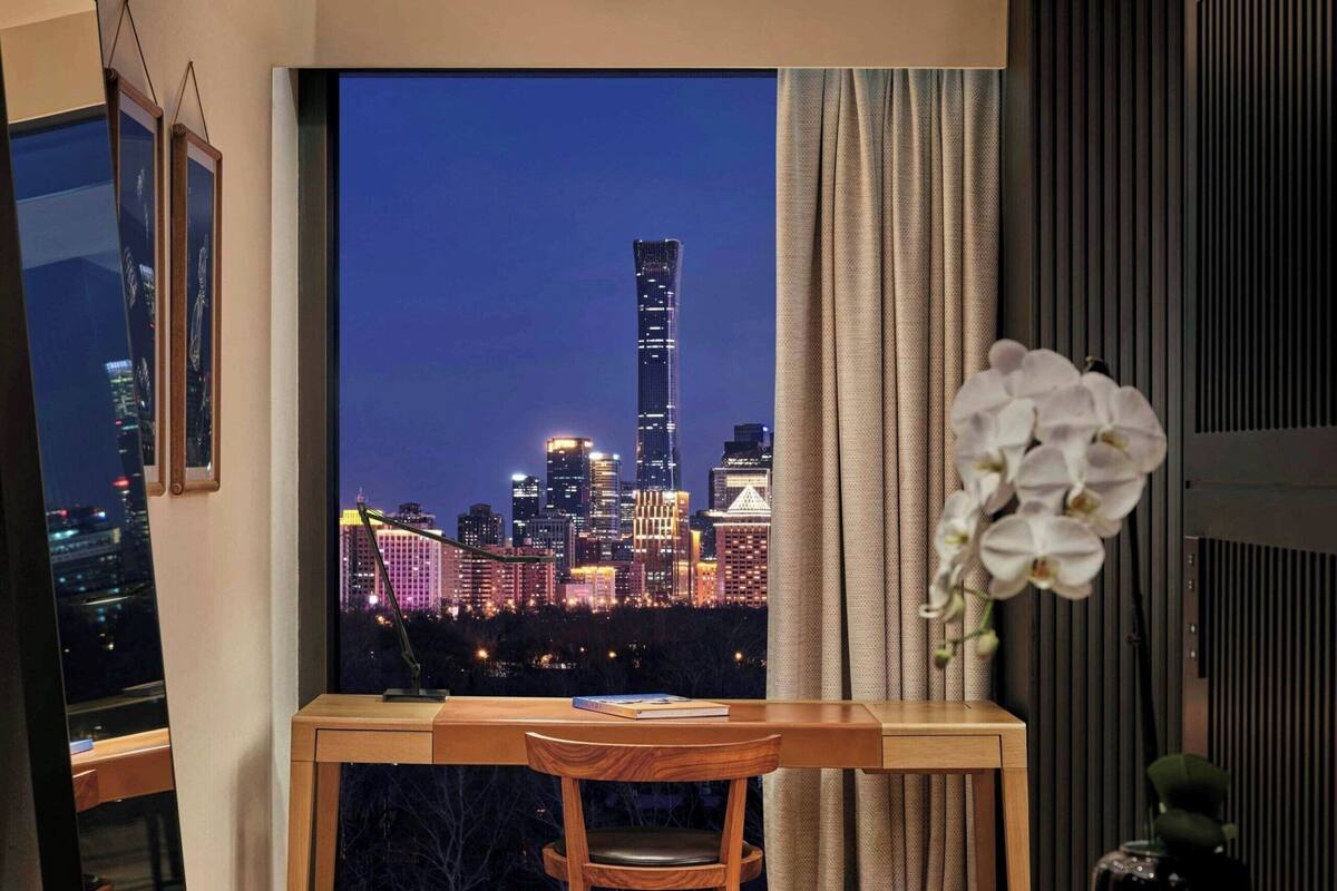 A modern room with a wooden floor, a desk, a chair, a large window with a city view, and a potted orchid.