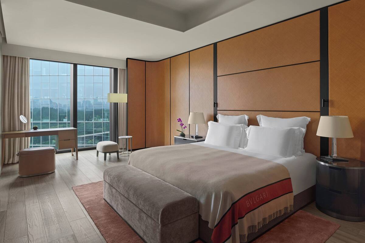A modern hotel room with a large bed, a desk, a chair, a small table, and a view of the city through large windows.