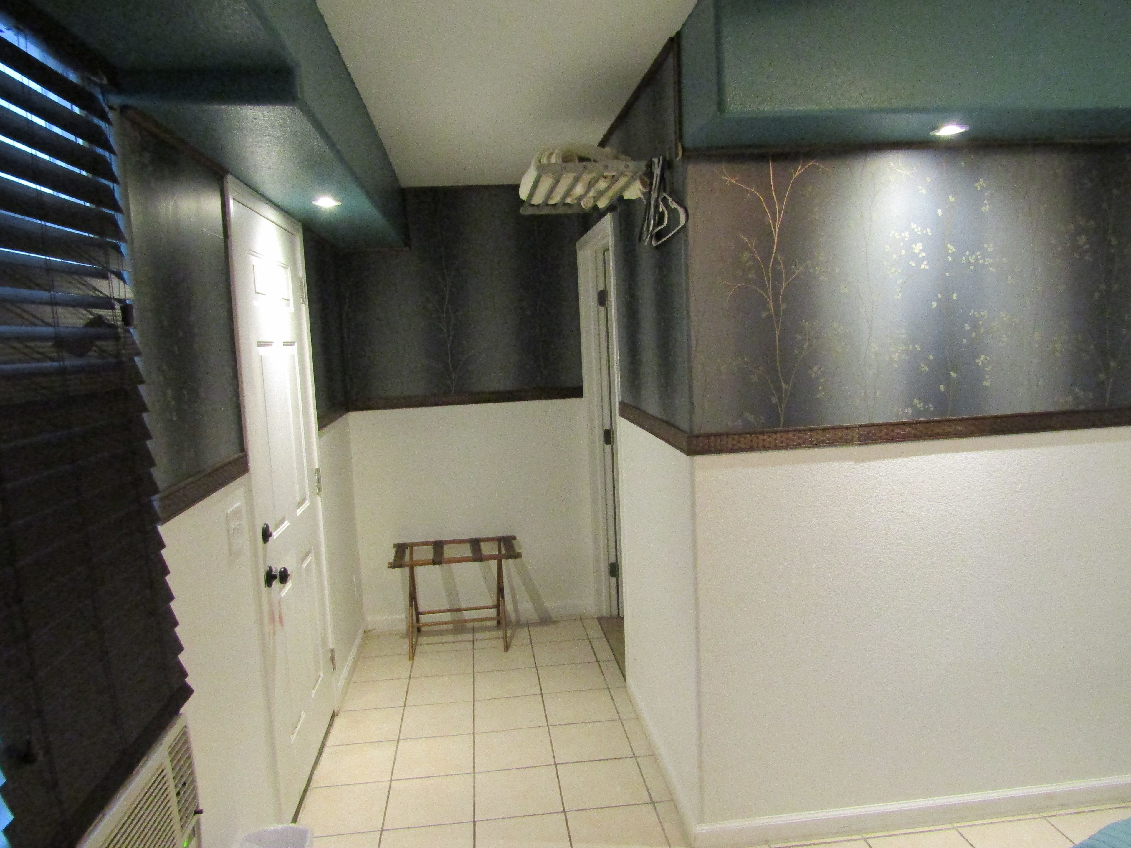 A narrow hallway with a tiled floor, a small wooden bench, a door, and a wall with a dark section and a lighter section.
