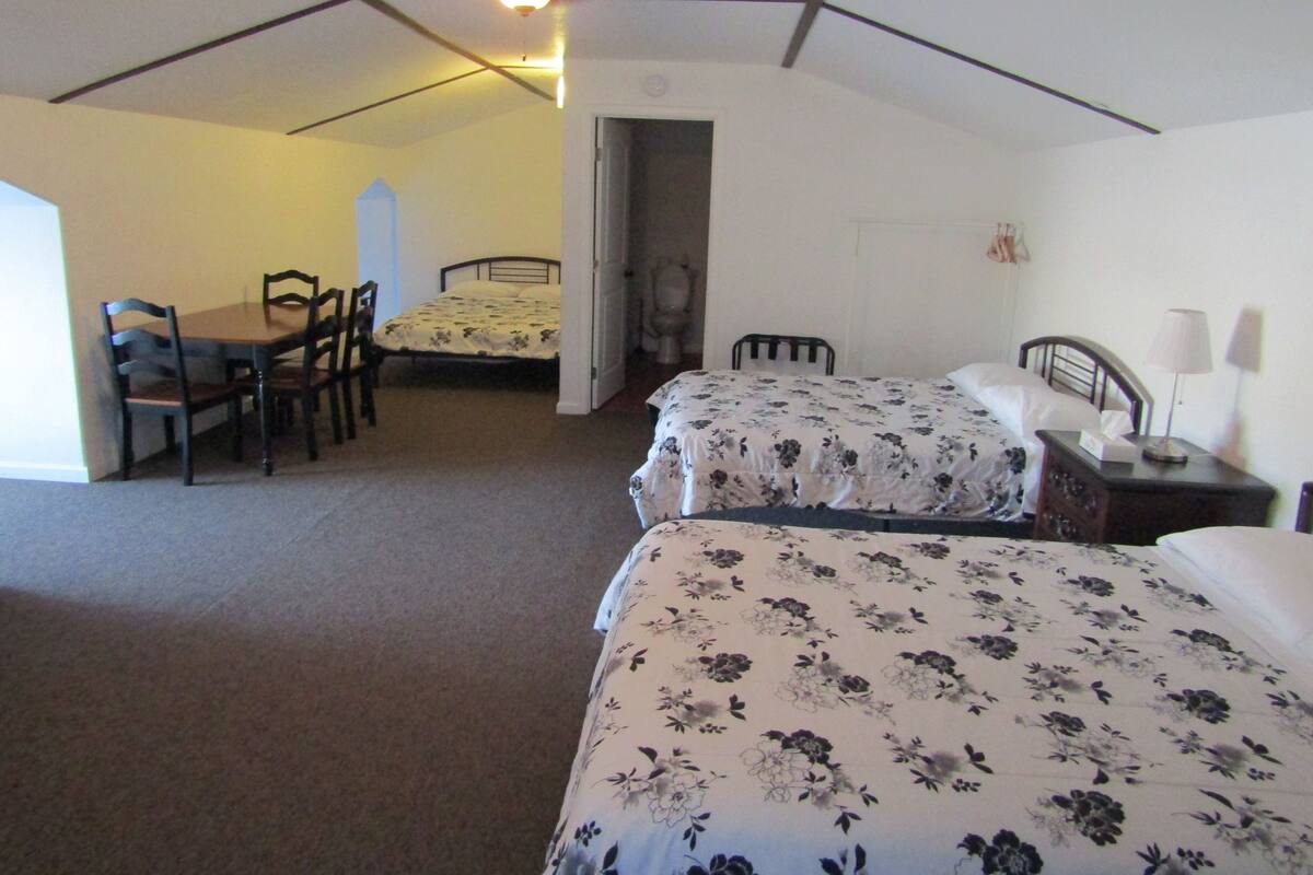A room with two beds, a dining table with chairs, a ceiling fan, and a door leading to another room.