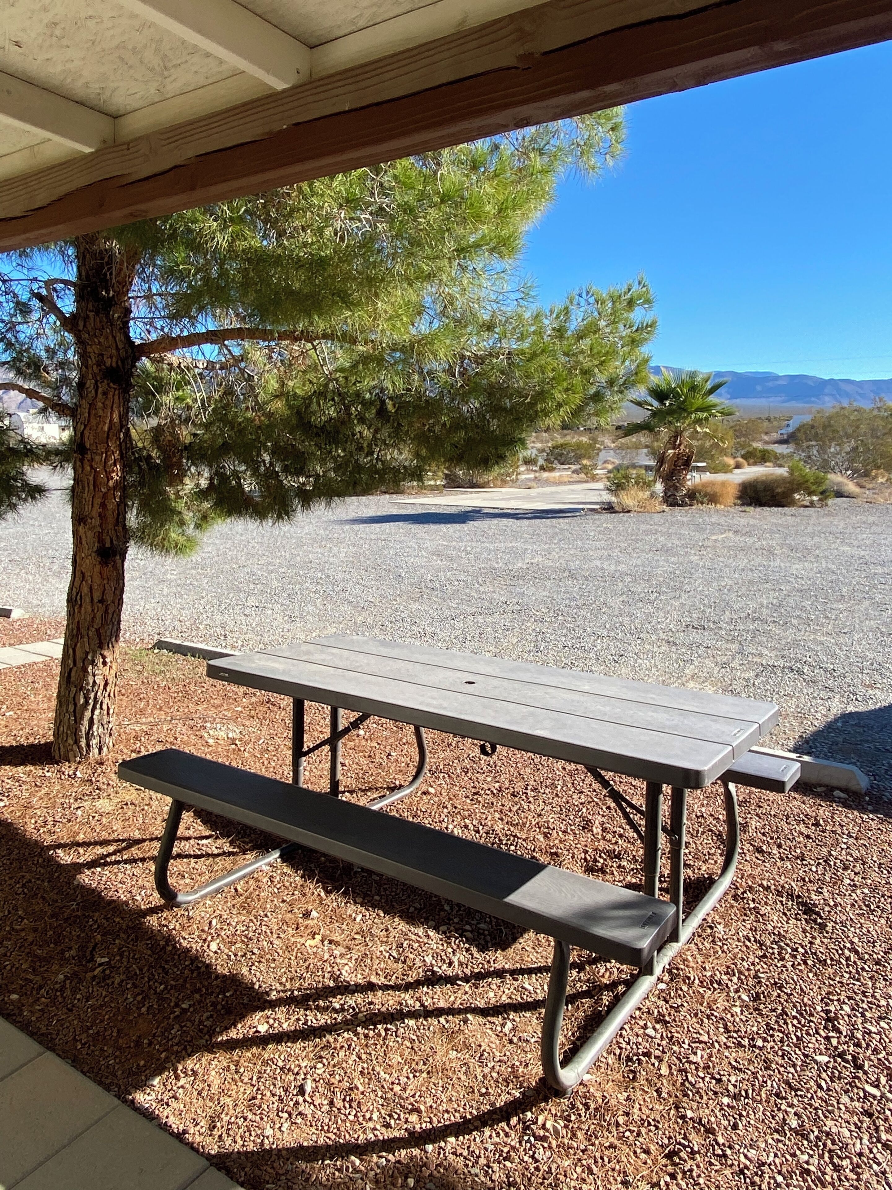 A picnic table under a wooden shelter with a gravel ground and trees in the background.