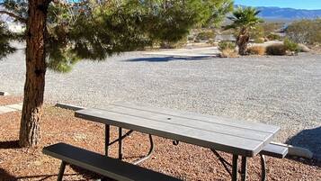 A picnic table under a wooden shelter with a gravel ground and trees in the background.