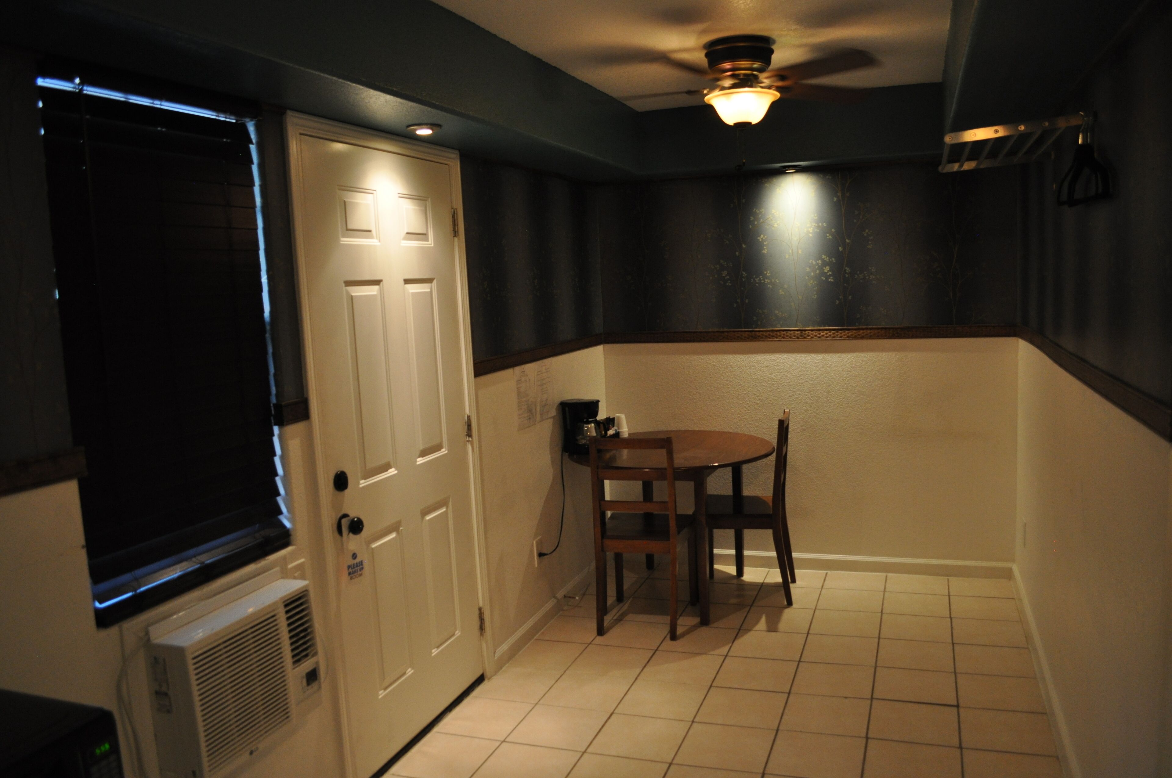A small kitchen with a white door, a table with two chairs, a ceiling fan, and a window with blinds.